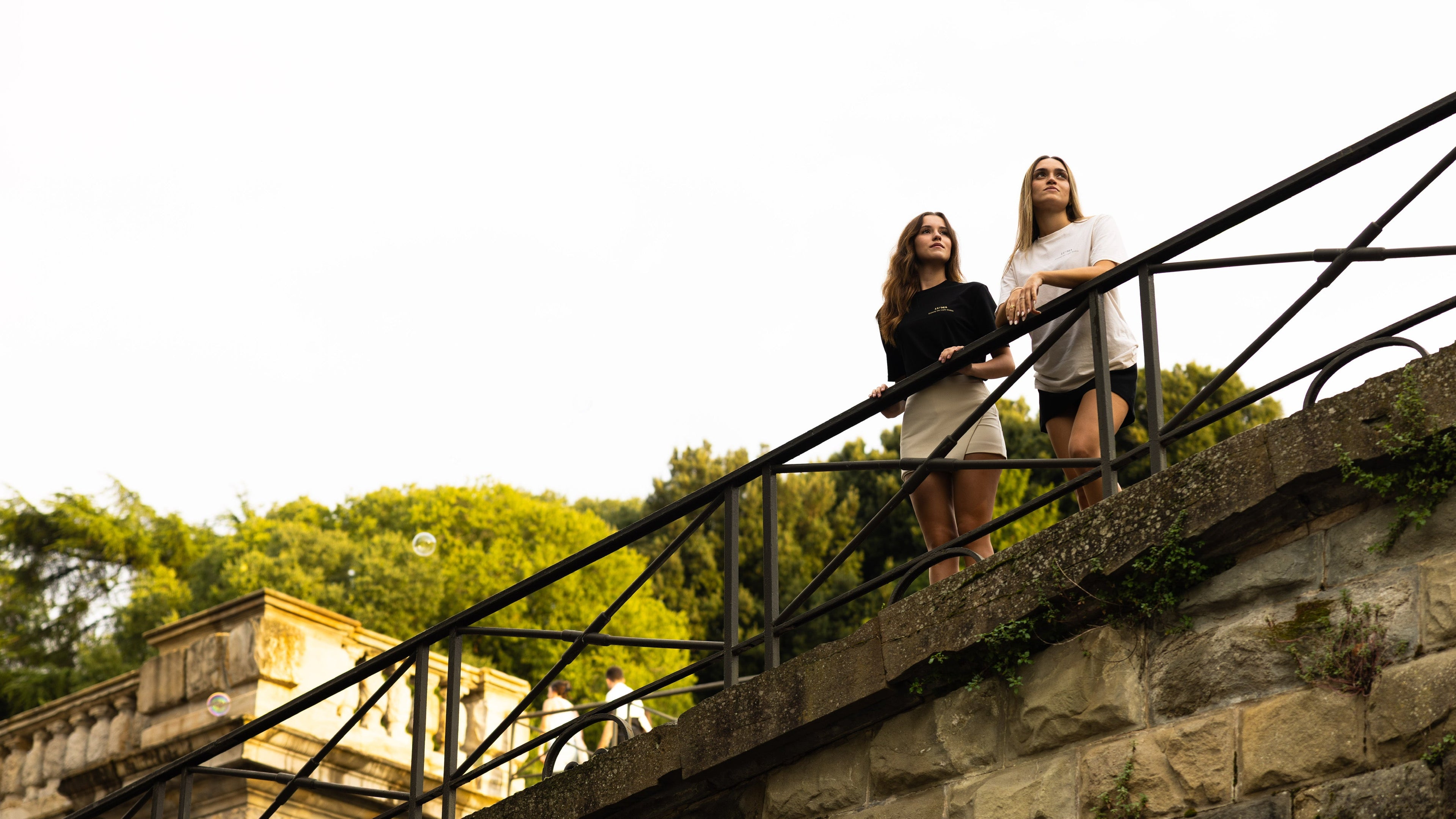 Two women standing on a stone staircase with greenery in the background wearing LU'MA clack and white t-shirts